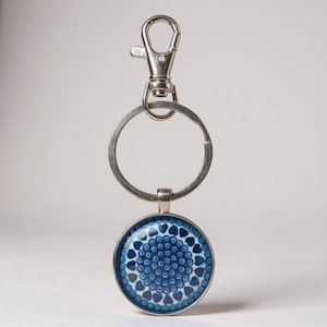 May include: A silver-toned keychain with a round pendant featuring a blue heart pattern. The keychain includes a split ring and a lobster clasp for easy attachment to keys or bags. The pendant has a silver border.
