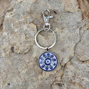May include: A silver keychain with a round pendant featuring a blue and white floral design. The pendant has a silver border and is attached to a split ring and a lobster clasp.