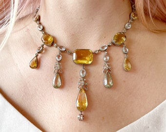 May include: A vintage necklace featuring a large, rectangular yellow gemstone at the center, surrounded by smaller, round and teardrop-shaped yellow and clear gemstones. The necklace has a silver-colored metal setting and a delicate, ornate design.