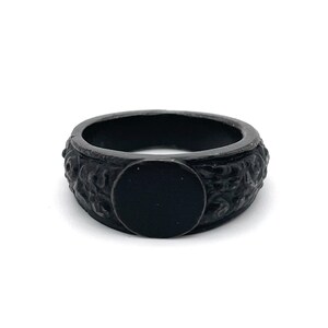 Puede incluir: Un anillo negro con una banda texturizada y una cara plana circular. El anillo tiene un acabado oscuro y mate y parece estar hecho de metal. La banda presenta un patr&oacute;n detallado, posiblemente floral.