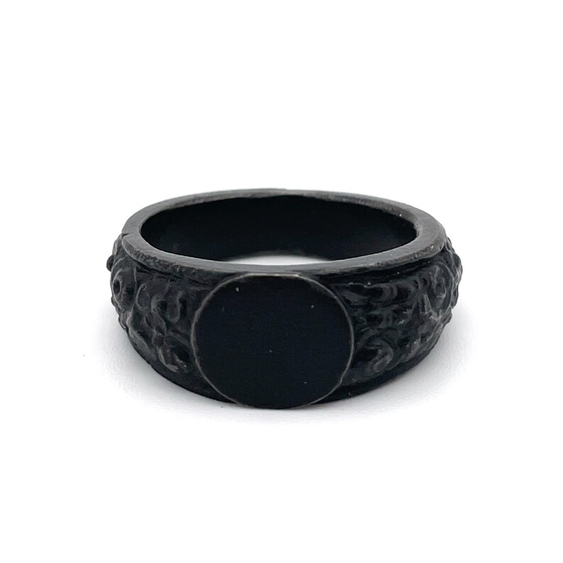 Puede incluir: Un anillo negro con una banda texturizada y una cara plana circular. El anillo tiene un acabado oscuro y mate y parece estar hecho de metal. La banda presenta un patr&oacute;n detallado, posiblemente floral.