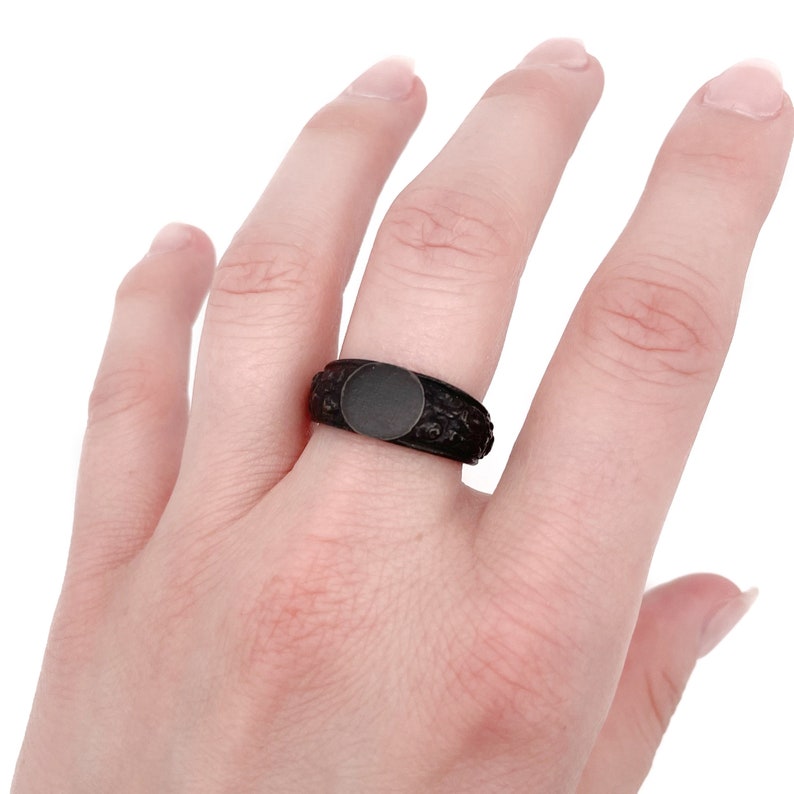 Puede incluir: Un anillo de sello negro con una cara redonda negra mate. La banda presenta un patr&oacute;n detallado, similar a flores. El anillo se lleva en un dedo, mostrando su dise&ntilde;o y textura. El color oscuro del anillo contrasta con el tono de la piel.