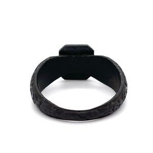 May include: A black ring with a square-cut, dark gemstone. The band has a textured, patterned design. The ring is set against a white background, highlighting its dark color and intricate details. A piece of jewelry.