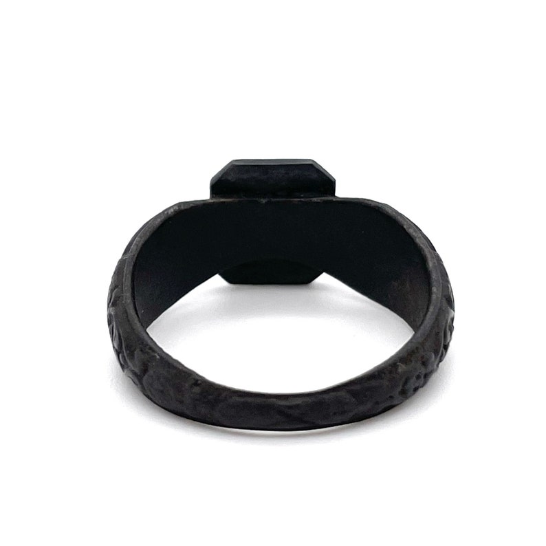 May include: A black ring with a square-cut, dark gemstone. The band has a textured, patterned design. The ring is set against a white background, highlighting its dark color and intricate details. A piece of jewelry.