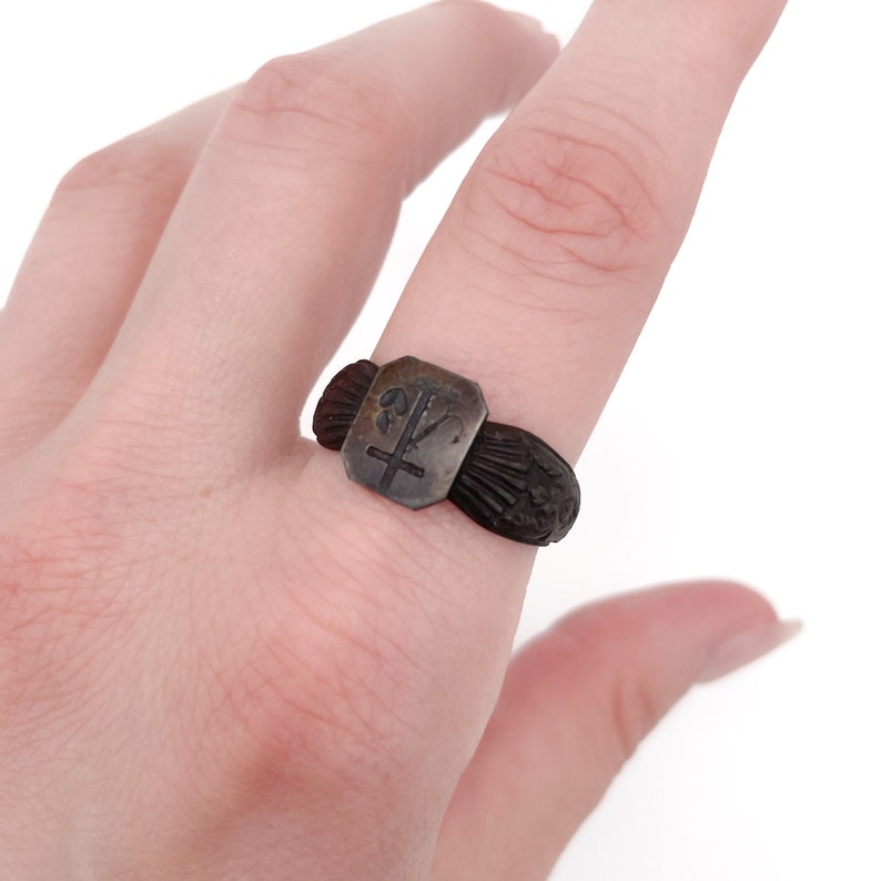 May include: A dark, antique-style ring with a rectangular face featuring a cross and floral design. The ring has a textured band and decorative elements on either side of the face. The ring is worn on a finger.