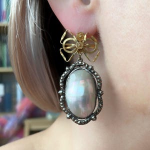May include: A gold-toned bow-shaped earring top with a decorative center piece, connected to a large, oval-shaped pearl pendant. The pearl is surrounded by a dark, ornate frame. The earring is a statement piece.