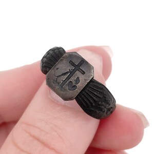 May include: A small, antique silver ring with a dark, textured band. The ring features a rectangular silver centerpiece with a cross, a heart, and other symbols. The ring is held by a person.