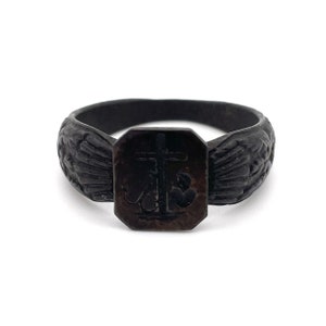 May include: A dark, antique-style ring with a rectangular centerpiece featuring a cross and anchor design. The ring's band has a textured, carved pattern. The ring is a dark, possibly oxidized, metal.