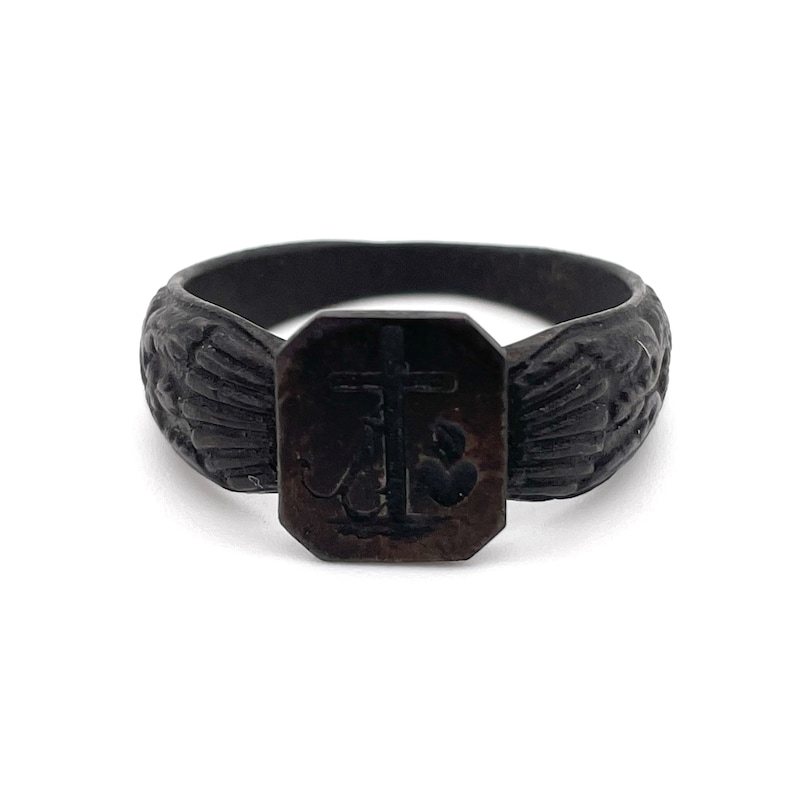 May include: A dark, antique-style ring with a rectangular centerpiece featuring a cross and anchor design. The ring's band has a textured, carved pattern. The ring is a dark, possibly oxidized, metal.