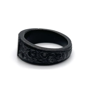 Puede incluir: Un anillo negro con una cara rectangular y un intrincado dise&ntilde;o floral alrededor de la banda. El anillo, hecho de un material oscuro y mate, se presenta sobre un fondo blanco.