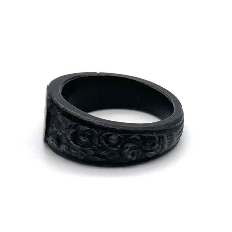 Puede incluir: Un anillo negro con una cara rectangular y un intrincado dise&ntilde;o floral alrededor de la banda. El anillo, hecho de un material oscuro y mate, se presenta sobre un fondo blanco.