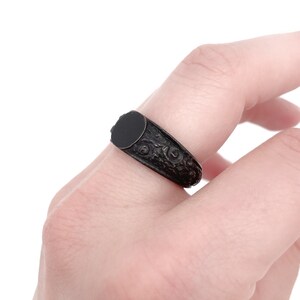 Puede incluir: Anillo de sello negro con una parte superior redonda y plana y detalles florales ornamentados en la banda. El anillo se lleva en un dedo, mostrando su acabado oscuro y mate y su dise&ntilde;o cl&aacute;sico. Una joya.