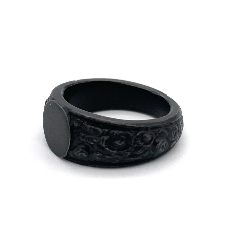 Puede incluir: Un anillo negro con acabado mate. El anillo presenta un patr&oacute;n floral en relieve alrededor de la banda y una cara plana de forma ovalada. El anillo es de color oscuro s&oacute;lido y el dise&ntilde;o es detallado.
