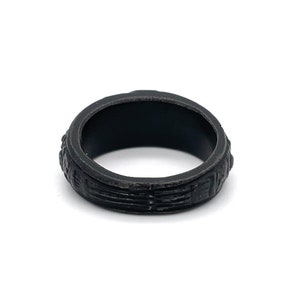 Puede incluir: Un anillo negro con una superficie texturizada. El anillo tiene una banda de l&iacute;neas horizontales y elementos decorativos en los bordes. El anillo est&aacute; sobre un fondo blanco.