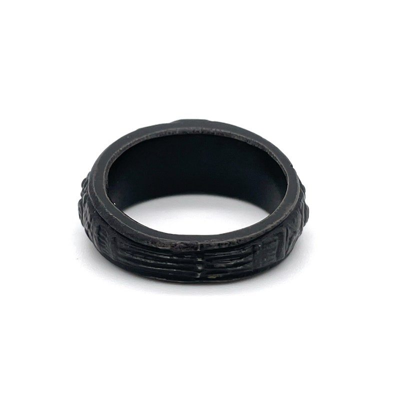 Puede incluir: Un anillo negro con una superficie texturizada. El anillo tiene una banda de l&iacute;neas horizontales y elementos decorativos en los bordes. El anillo est&aacute; sobre un fondo blanco.