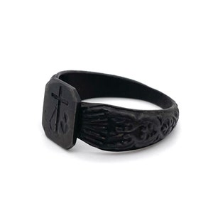 May include: A black ring with a rectangular face featuring a cross and an anchor design. The band has an ornate, carved pattern. The ring is set against a white background.