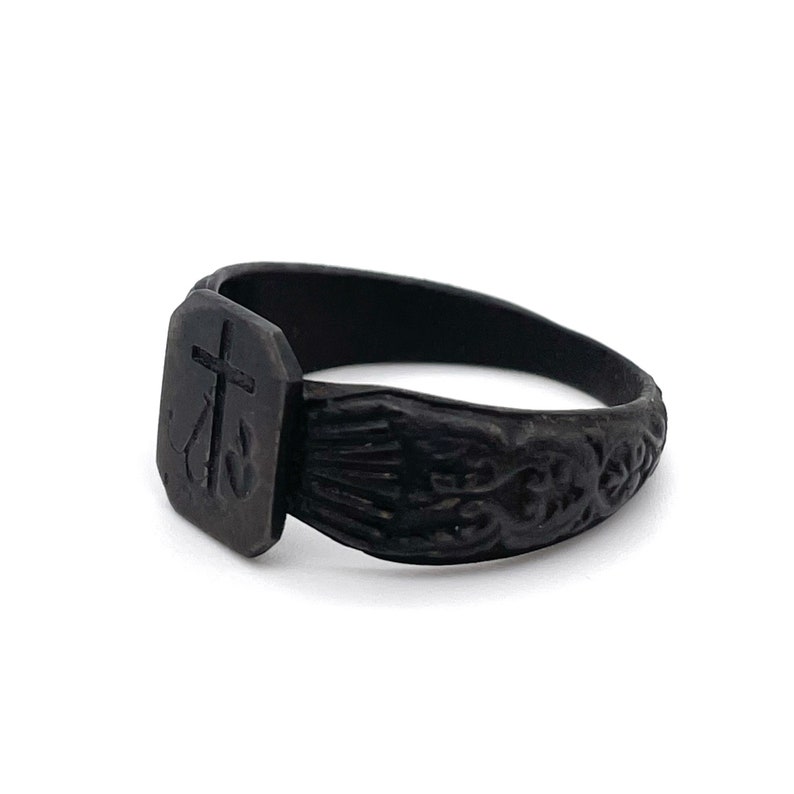 May include: A black ring with a rectangular face featuring a cross and an anchor design. The band has an ornate, carved pattern. The ring is set against a white background.