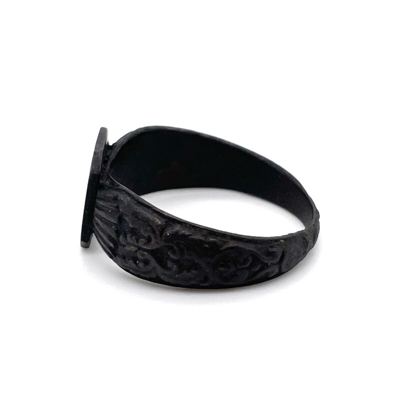 May include: A black ring with a rectangular face and ornate, swirling patterns on the band. The ring is made of a dark metal and has a vintage or antique aesthetic. The ring is shown on a white background.