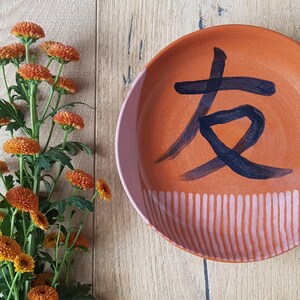 May include: A ceramic plate with a pink, orange, and teal glaze. The plate has a black painted Chinese character for "friend" on it.