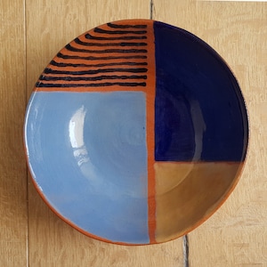 Op de afbeelding: Een keramische kom met een geometrisch ontwerp. De kom is verdeeld in vier secties, elk geverfd in een andere kleur: blauw, lichtblauw, oranje en bruin. De bovenste sectie heeft een patroon van zwarte lijnen op een oranje achtergrond.