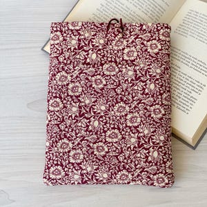 Può includere: Una custodia per libri con motivo floreale bordeaux e crema con chiusura a coulisse. La custodia è rettangolare e poggia su un libro aperto. Il motivo floreale presenta fiori e foglie dettagliate.