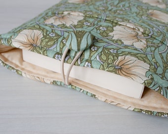 Könnte beinhalten: Ein Buchcover aus beigefarbenem Stoff mit einem floralen Muster in Grün-, Weiß- und Brauntönen. Der Stoff hat einen dezenten Glanz und die Blumen sind stilisiert und filigran. Das Buchcover ist geschlossen und hat zwei beigefarbene Stoffbänder.