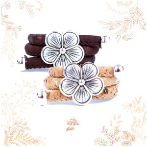 Puede incluir: Dos pulseras de corcho con abalorios de flores plateadas. Las pulseras están envueltas en corcho y tienen una cuenta plateada al final. Los abalorios de flores son detallados y tienen un aspecto vintage.