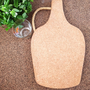 May include: A light brown cork serving board with a rope handle. A clear glass with a floral design and fresh green herbs are next to the board. The board is approximately 15 inches long.