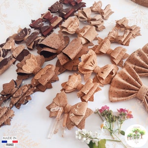 May include: A collection of cork hair bows in various shapes and sizes. The bows are made from natural cork and have a rustic, earthy look. Some of the bows feature gold accents. The bows are displayed on a white surface with a floral background.