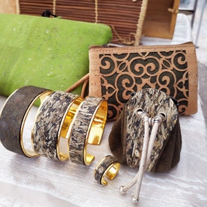 May include: Four cork and gold bracelets with a small cork and gold ring. The bracelets are stacked on top of each other. There is a small cork bag with a drawstring closure and a cork purse with a green and brown patterned design.