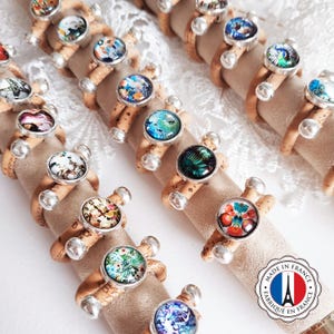 Puede incluir: Una colección de anillos de corcho y plata con inserciones de vidrio de colores. Cada anillo presenta un diseño diferente. Los anillos se muestran sobre un fondo blanco con una etiqueta que dice "Made in France - Fabriqué en France".
