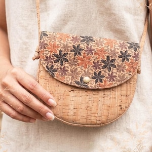 May include: A semi-circular crossbody bag made of natural cork material. The flap features a repeating floral pattern in shades of brown, orange, and dark grey, secured by a single light-coloured circular button closure and a thin shoulder strap.