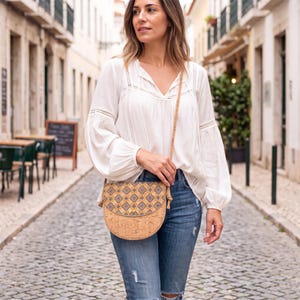 May include: A crossbody bag made of natural cork material featuring a rounded bottom and a flap with a blue and gold geometric pattern. The bag has a thin cork strap and is worn over a white long-sleeved blouse and blue distressed denim jeans.