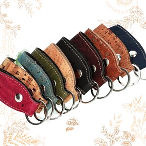 Op de afbeelding: Een verzameling sleutelhangers in verschillende kleuren, waaronder rood, blauw, groen en bruin. Elke sleutelhanger heeft een kurken band met een metalen ring en een zilveren drukknoopsluiting. De sleutelhangers zijn diagonaal gerangschikt.