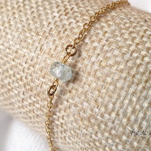 Op de afbeelding: Een delicate gouden kettingarmband met een enkele lichtblauwe aquamarijn kraal. De armband wordt getoond op een jute achtergrond.