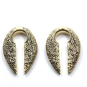 Könnte beinhalten: Ein Paar goldfarbene Ohrdehner aus Messing mit aufwendigen floralen Designs. Die Dehnungen sind tropfenförmig mit einer Schlüssellochaussparung.