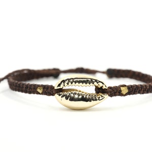 Könnte beinhalten: Ein braunes geflochtenes Armband mit einem goldenen Kaurischalen-Anhänger und goldenen Akzenten.