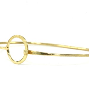 Può includere: Un braccialetto rigido in oro con un grande designo circolare. Il cerchio è aperto e il braccialetto è sottile.