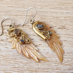 Op de afbeelding: Een paar gouden en zilveren oorbellen met een gesneden schelpvleugelontwerp. Elke vleugel is versierd met een blauwe edelsteen en ingewikkelde gouden filigraan.