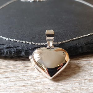 Könnte beinhalten: Ein polierter herzförmiger Medaillon-Anhänger aus Silber, der an einer zarten Silberkette hängt. Das Medaillon hat eine glatte, reflektierende Oberfläche und eine einfache Öse an der Oberseite. Der Text MELATI jewelry design ist unten links sichtbar.