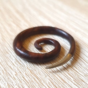 May include: A dark brown wooden spiral ear gauge with a silver metal tip. The gauge is lying on a light wood surface.