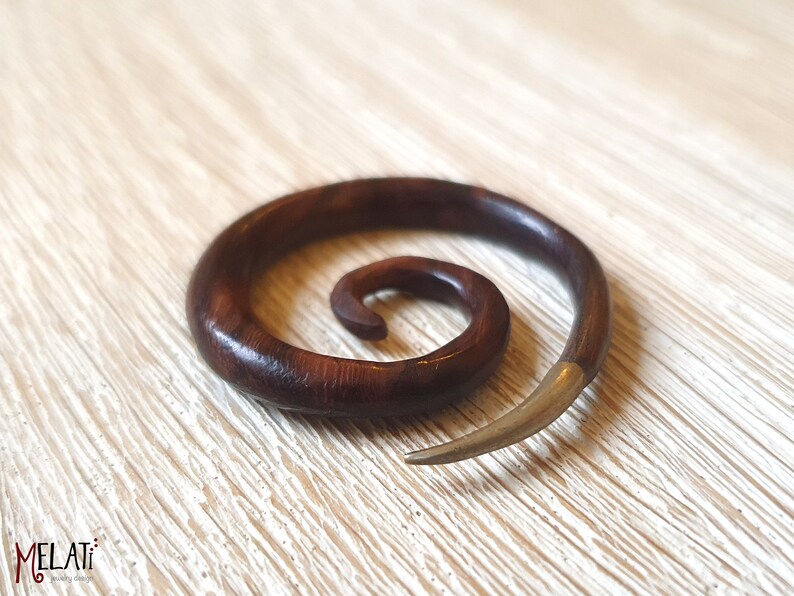 May include: A dark brown wooden spiral ear gauge with a silver metal tip. The gauge is lying on a light wood surface.