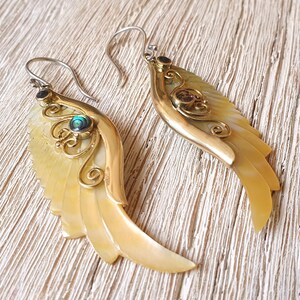 Op de afbeelding: Een paar gouden en witte schelp oorbellen met een groene edelsteen accent. De oorbellen zijn gevormd als vleugels en hebben ingewikkelde details.