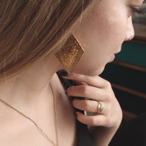 May include: A woman wearing a black top with a gold chain necklace featuring a turquoise stone pendant. She is also wearing a gold ring with a turquoise stone and a pair of large gold earrings with a geometric design.