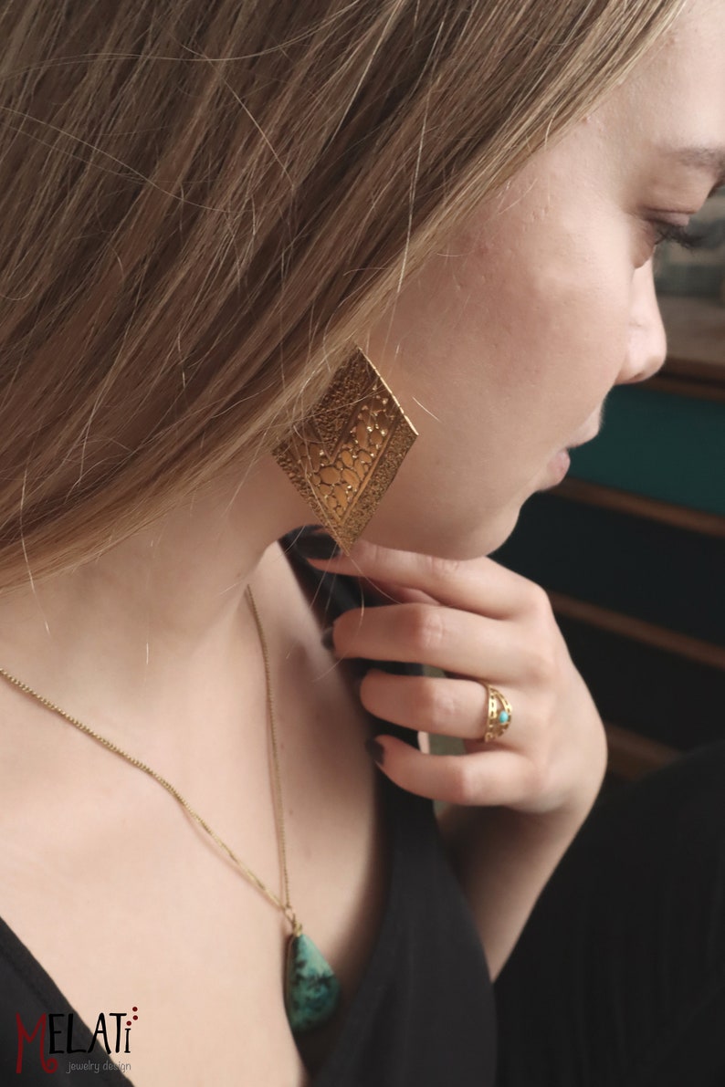 May include: A woman wearing a black top with a gold chain necklace featuring a turquoise stone pendant. She is also wearing a gold ring with a turquoise stone and a pair of large gold earrings with a geometric design.