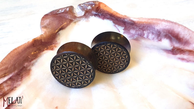 May include: A pair of wooden ear gauges with a geometric pattern. The gauges are brown with a black geometric design.