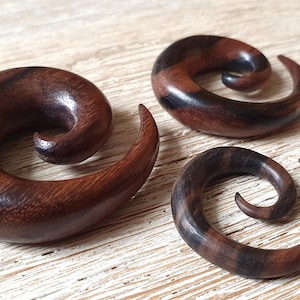 Könnte beinhalten: Drei spiralförmige Ohrstecker aus Holz in verschiedenen Größen. Die Stecker sind aus dunkelbraunem Holz mit natürlicher Maserung.