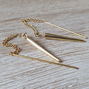 Könnte beinhalten: Goldfarbene Ohrringe mit Ketten- und Stangendesign. Die Ohrringe haben einen langen, schlanken Stab, der an einer Kette hängt, mit einem konischen Element. Der Schmuck wird auf einer hellen Holzoberfläche präsentiert.
