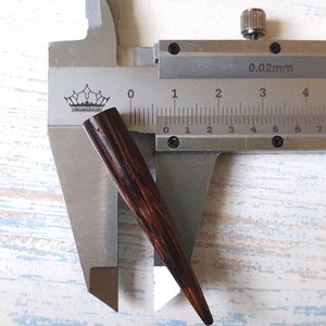 May include: A dark brown, tapered wooden spike or jewelry component positioned within a metal vernier caliper. The caliper displays a measurement of approximately 0.5 inches in width at the widest point of the dark, grain-textured object.