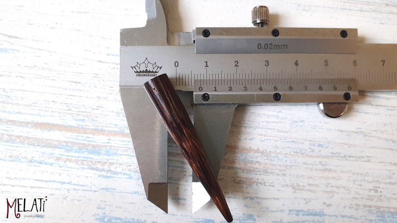 May include: A dark brown, tapered wooden spike or jewelry component positioned within a metal vernier caliper. The caliper displays a measurement of approximately 0.5 inches in width at the widest point of the dark, grain-textured object.